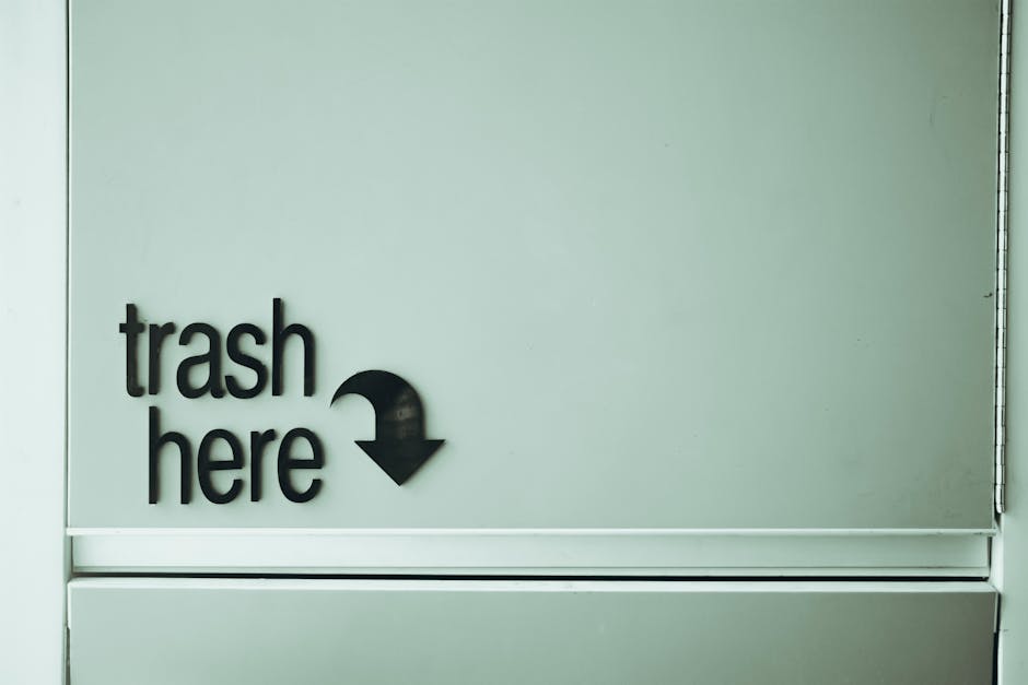 A close-up view of a pale green or light grey wall panel, featuring a black, three-dimensional sign that reads 'trash here' with a downward-pointing arrow. The sign is mounted on the wall, with the te
