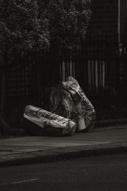 Three large black plastic garbage bags, visibly crumpled and protruding at their openings, are placed on a sidewalk curb near a black metal fence with vertical bars. The bags contain mixed waste, and 
