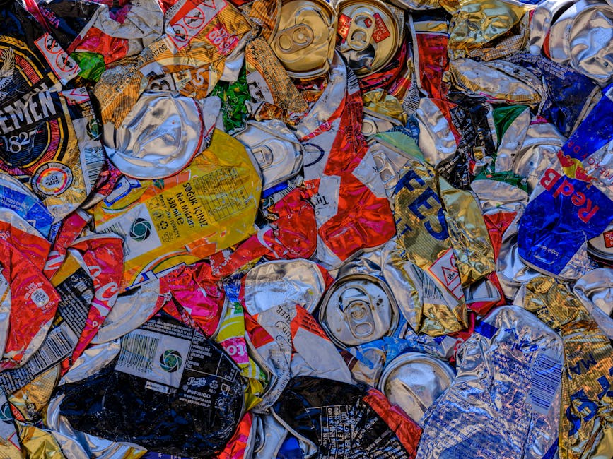 Close-up view of a large pile of crushed and crumpled aluminium beverage cans, predominantly silver with some red, gold, blue, and black sections, scattered randomly on a surface. The cans display var