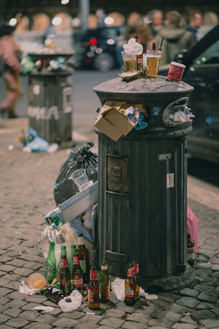 Close-up view of a large pile of crushed and crumpled aluminium beverage cans, predominantly silver with some red, gold, blue, and black sections, scattered randomly on a surface. The cans display var